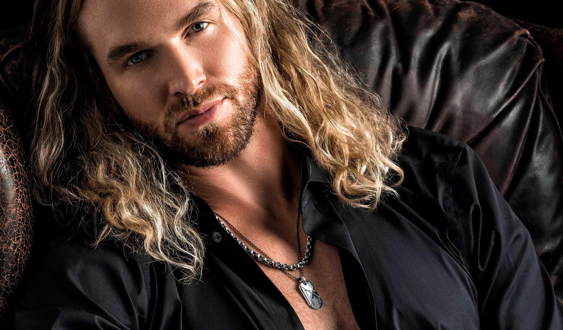 Long blond haired man sitting in leather couch with chain around beck